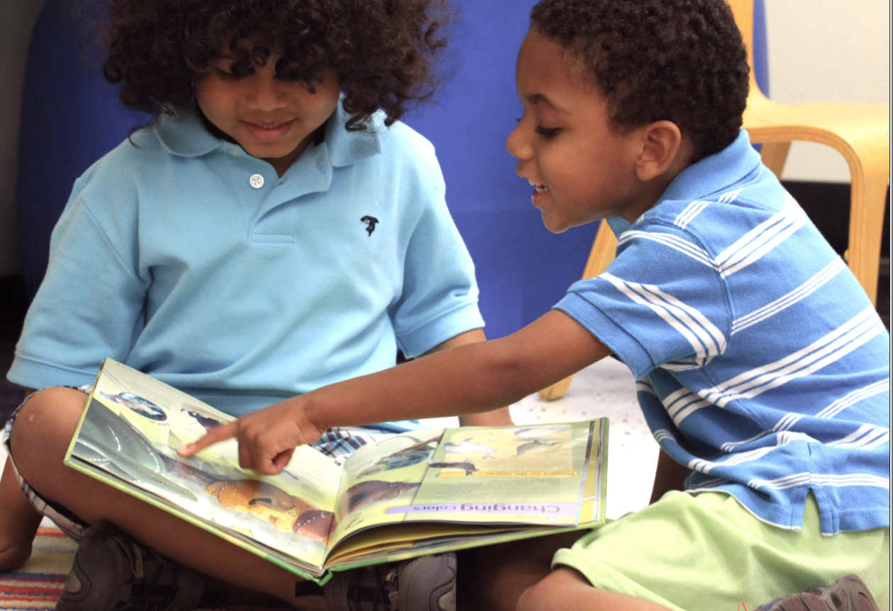 Two Black schoolchildren read a book together in a classroom setting.