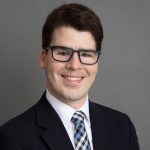 An image of a White man with brown hair wearing glasses, a blue, black, and white plaid tie and a dark suit.