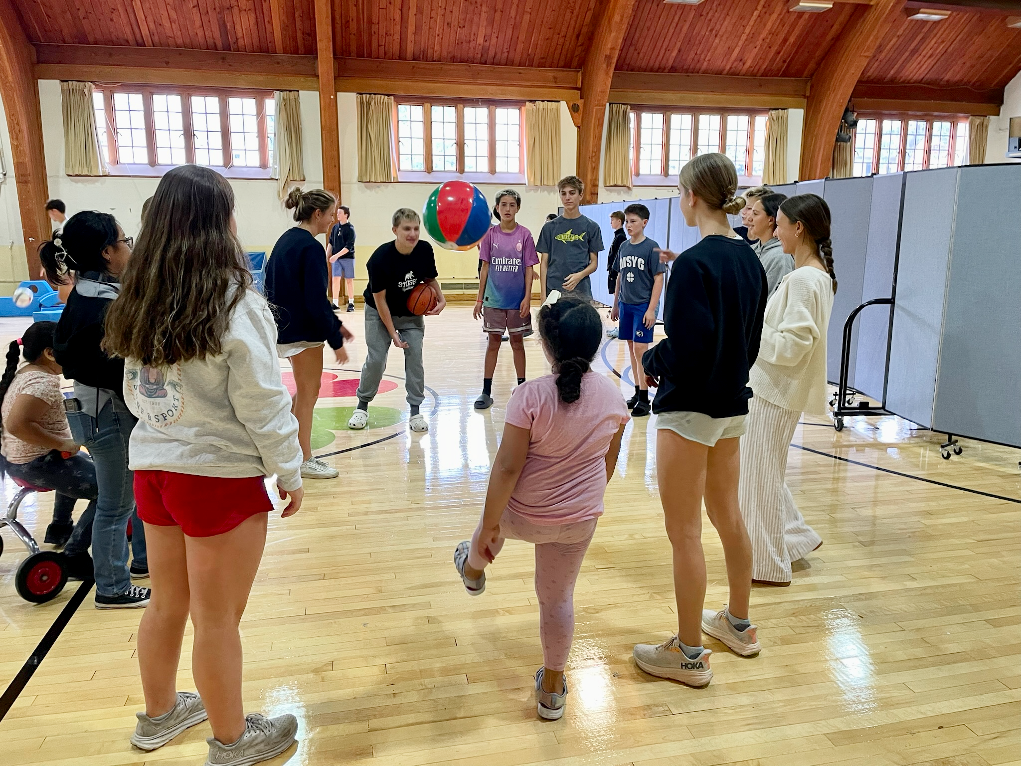 A group of students ranging in age from early childhood to high school stand in a circle inside a gymnasium playing a game with a beach ball.