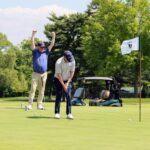 A golfer raises his hands above his head in victory while another golfer sinks a putt.