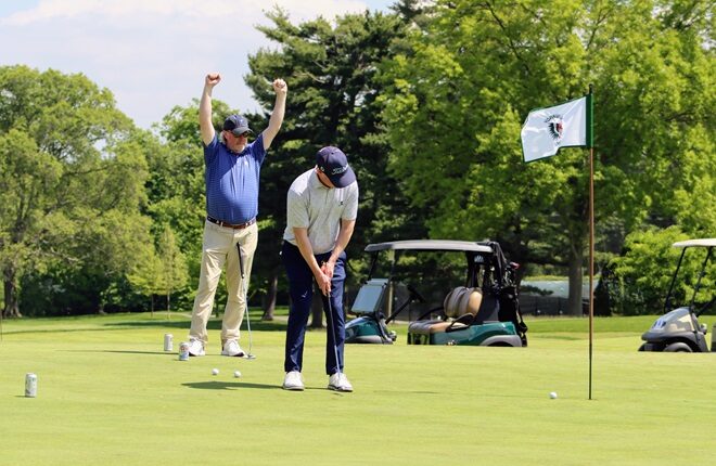 A golfer raises his hands above his head in victory while another golfer sinks a putt.