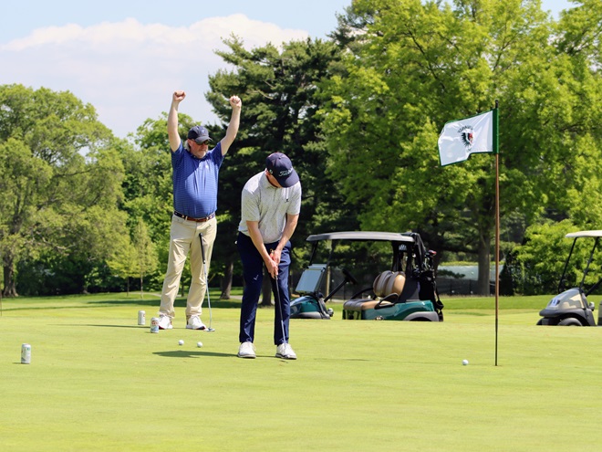 A golfer raises his hands above his head in victory while another golfer sinks a putt.