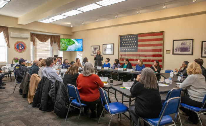 A group of 30 professionals and board members sit at a board room table during a conference.