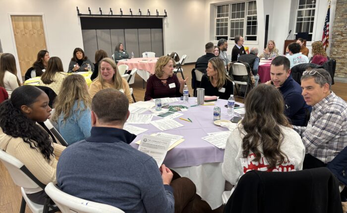 A large room full of adults and teens sit at round tables and talk with each other.