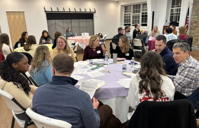 A large room full of adults and teens sit at round tables and talk with each other.