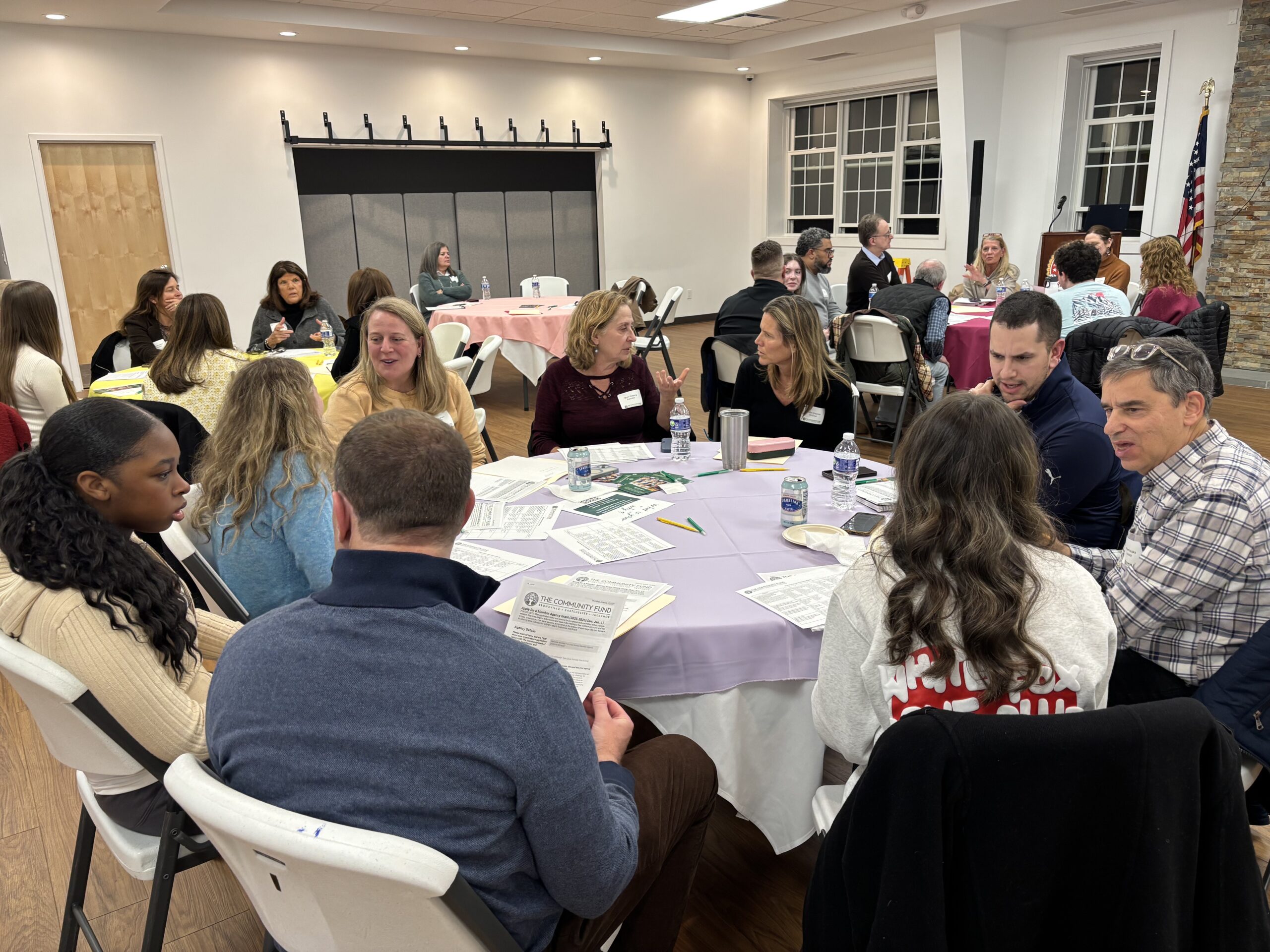 A large room full of adults and teens sit at round tables and talk with each other.