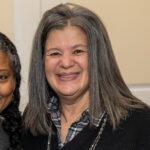 Photo of White woman with straight salt-and-pepper hair wearing a black top and necklace.