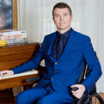 Photo of a White male with brown hair sitting in a wheelchair wearing a blue suit.