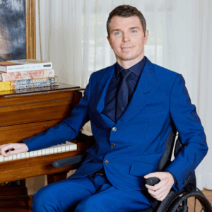 Photo of a White male with brown hair sitting in a wheelchair wearing a blue suit.