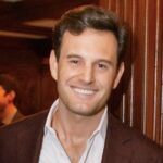 A photo of a White man with brown hair wearing a brown blazer and a white buttondown shirt.