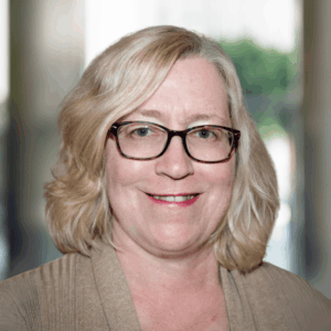 Headshot of a White woman with blonde hair wearing glasses and a beige top.