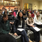 Photo of an audience of teen volunteers seated in chairs. Their keynote speaker stands at the right of the frame.
