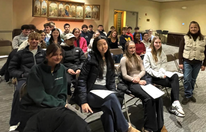 Photo of an audience of teen volunteers seated in chairs. Their keynote speaker stands at the right of the frame.